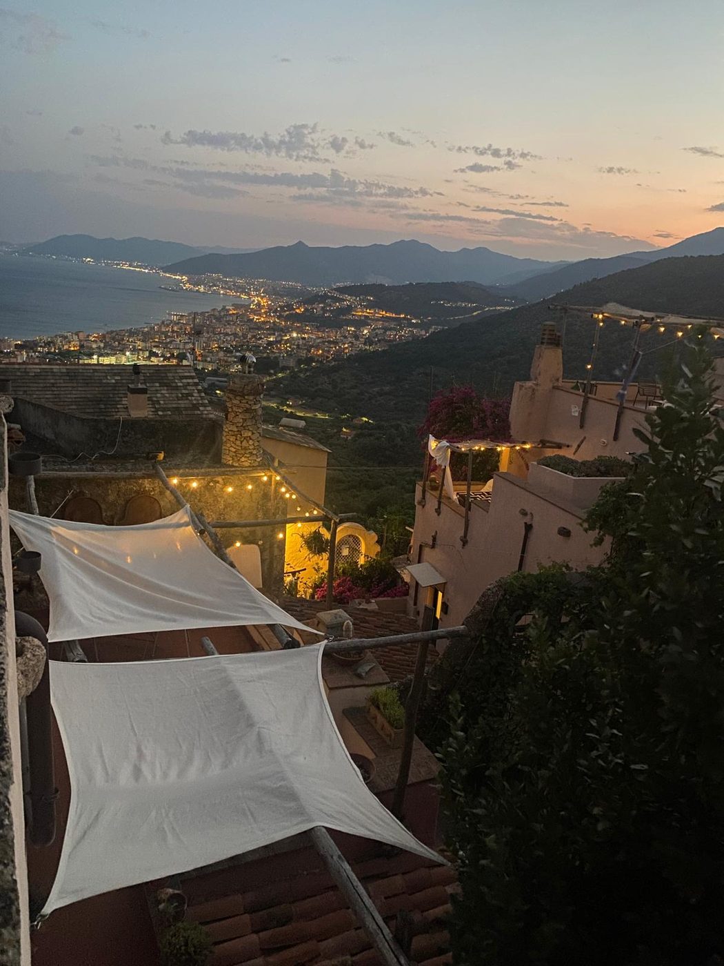 Dusk view over the stone rooftops of Verezzi, Liguria, with string lights and the Riviera di Ponente coastline beyond